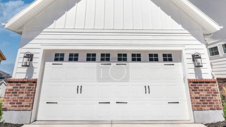 Téléchargez les photos : Panorama Nuages blancs gonflés Extérieur d'un garage avec parement en bois blanc et briques rouges. Il y a deux portes de garage blanches charnières latérales avec panneaux de fenêtre et une vue sur le quartier avec pelouse et maison inachevée sur la droite. - en image libre de droit