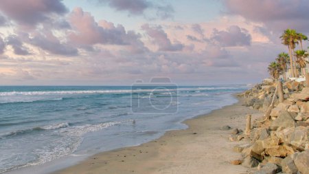 Foto de Panorama Nubes hinchadas al atardecer Playa vacía con rompeolas natural en Oceanside, California. Hay un mar a la izquierda con una orilla arenosa y una vista de palmeras a la izquierda fuera de los edificios. - Imagen libre de derechos