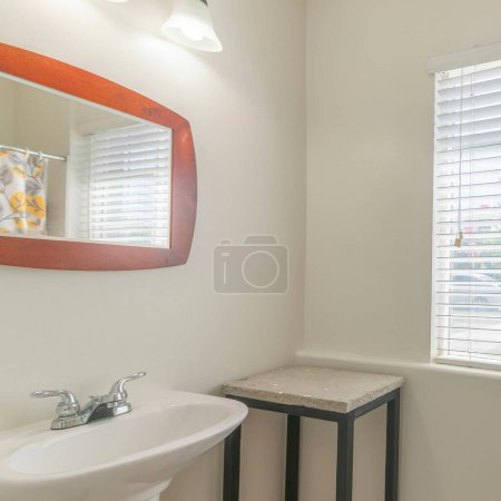 Photo for Square Bathroom interior with table and pedestal sink with framed mirror. Small bathroom with printed shower curtain on the left beside the toilet near the window with blinds. - Royalty Free Image