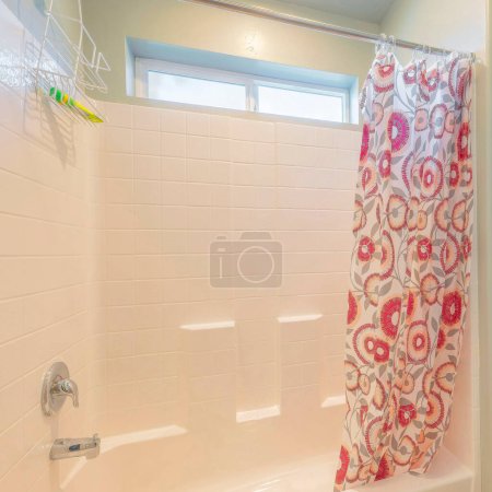 Quadratisch Kleines Badezimmer mit hängendem Duschkopf Caddy. Links neben der Wannendusche befindet sich eine Toilette mit bedrucktem Duschvorhang und Fliesenmuster auf der Acrylwand am Fenster.