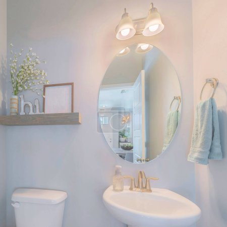 Photo for Square Powder room interior with decorations on the shelf. There is a wall mount toilet paper holder on the left near the toilet bowl beside the pedestal sink with oval mirror and hanging towel. - Royalty Free Image