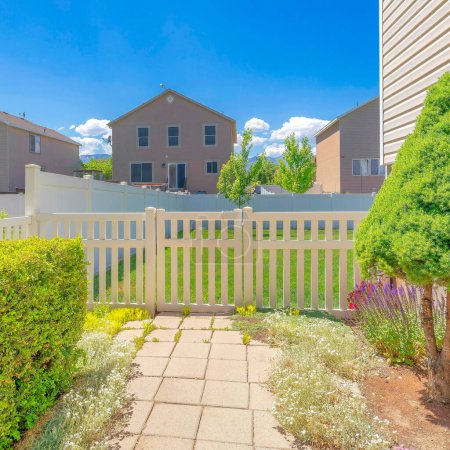 Photo for Square White entrance gate with fence of a backyard in Utah. There is a square cement tile floors of a path with shrubs on the side near the house on the left with vinyl wood siding leading to the backyard. - Royalty Free Image