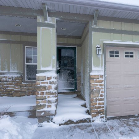 Photo for Square Exterior of a house with two garage doors and stone bricks siding. House with green and brown tone colors and a snow-covered driveway and doorsteps. - Royalty Free Image