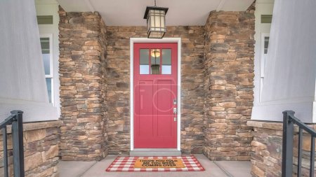 Panorama Exterior de una entrada frontal de una casa con paredes de piedra. Hay dos barandillas metálicas en el lateral que conducen a una puerta de hormigón con un felpudo grande debajo de la puerta delantera roja con paneles de vidrio.