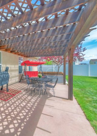 Téléchargez les photos : Terrasse arrière verticale avec toit en pergola en bois et barbecue couvert. Il y a une porte avec du verre et un ensemble de table et chaises sur le côté et une pelouse sur la droite. - en image libre de droit