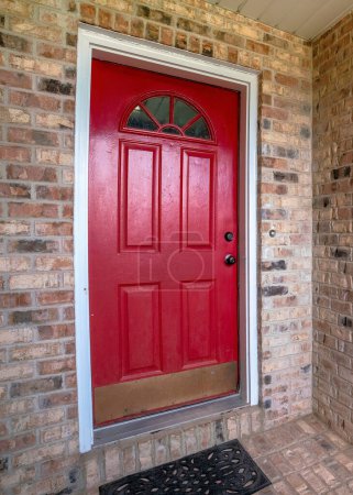 Photo for Vertical Entrance exterior of a house with red door and bricks. There is a red front door with arched glass panel near the sliding glass windows on the left across the shrubs. - Royalty Free Image