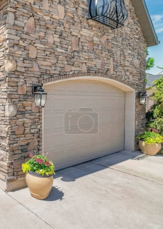 Vertical White puffy clouds Double arched sectional garage doors with potted plants outside. Three car garage exterior with stone veneer siding and arched window with railings.