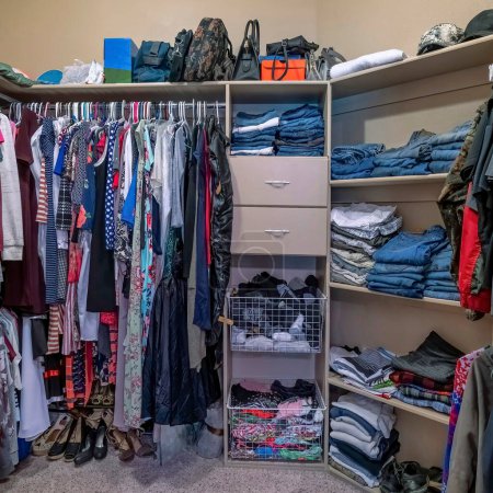 Photo for Square Full organized walk in closet with open wardrobe and carpeted flooring. There are clothes hanging on the metal rods and a stacked of denims on the front wall with shoes at the bottom. - Royalty Free Image