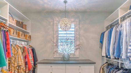 Photo for Panorama Large organized walk in closet with ball pendant lighting fixture. There are clothes hanging on both sides of the wall and a floor cabinet at the front with drawers and vase on top against the window. - Royalty Free Image