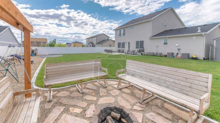 Photo for Panorama White puffy clouds Fenced backyard of a house with outdoor lounge and playground. There are wooden benches with a fire pit in the middle with woods beside the playground and a view of the house with patio. - Royalty Free Image