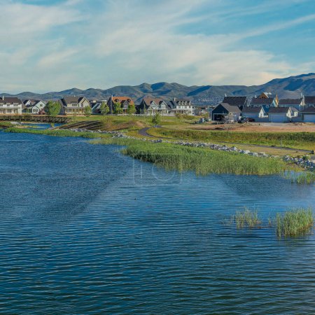 Photo for Square Whispy white clouds Oquirrh Lake at Daybreak in Utah with paved sidewalk on the side. There is a bridge over the lake and houses with different structures against the mountains at the background. - Royalty Free Image