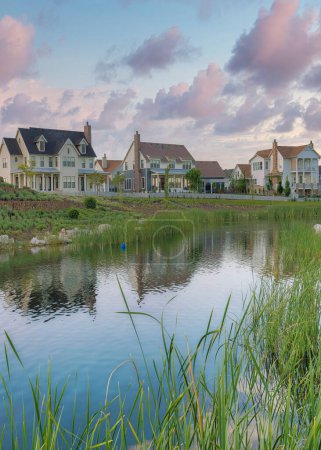 Photo for Vertical Puffy clouds at sunset Oquirrh Lake with a reflection of the residential houses at Daybreak, Utah. Lake waterfront with grass and rocks on the shore with a view of layered retaining walls on the left near the houses. - Royalty Free Image