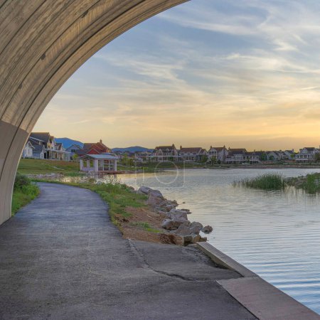 Photo for Square Whispy white clouds Under an arched bridge over Oquirrh Lake at Daybreak, Utah. There are two paths beside the lake with buoyancy balls and a view of residential houses against the sunset horizon sky at the back. - Royalty Free Image