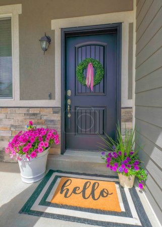 Vertical Black front door of a house with wreath and potted flowers at the front. Entrance exterior of a home with colorful doormat and a view of a window on the left with bench at the front.