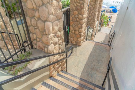 Photo for Stairs outside the fence and wall of the buildings at Carlsbad, San Diego, California. There is a fence with metal railings and stone posts on the left and a view of people at the beach. - Royalty Free Image