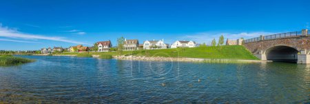 Photo for Panoramic view of Oquirrh Lake with grassy shore and stone bridge on the left. Lake waterfront of a residential area with large houses against the clear blue sky at the background. - Royalty Free Image