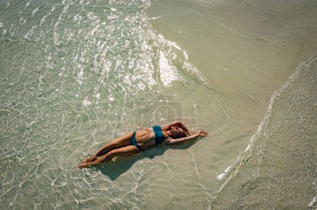 Foto de Una joven en bikini azul yace boca arriba sobre la arena cerca de las olas del mar azul. Vista desde arriba. La playa de las Maldivas, agua limpia y clara y arena amarilla - Imagen libre de derechos