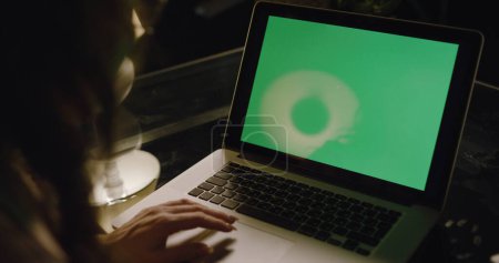 Foto de Sobre el hombro de una mujer de negocios que trabaja por la noche en una habitación con las luces apagadas en un ordenador en una mesa mirando una pantalla verde. Uso de ordenador portátil con pantalla portátil verde. - Imagen libre de derechos