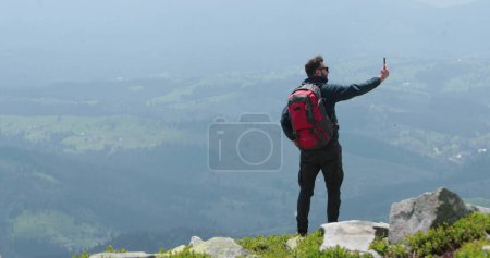 Foto de Un viajero en la cima de una montaña está hablando por teléfono, tomando fotos y teniendo un video chat. Concepto de comunicación en lugares remotos del mundo. - Imagen libre de derechos