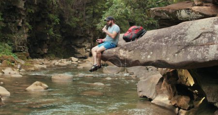 Foto de Un hombre-viajero solitario se sienta tranquilamente y descansa sobre una piedra que cuelga sobre el agua cerca del río contra el fondo del bosque y el lecho del río. - Imagen libre de derechos