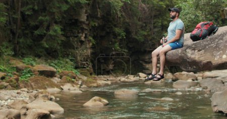 Foto de Un hombre-viajero solitario se sienta tranquilamente y descansa sobre una piedra que cuelga sobre el agua cerca del río contra el fondo del bosque y el lecho del río. - Imagen libre de derechos