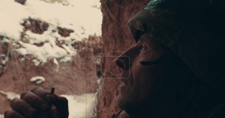 Foto de Un soldado anciano se sienta con una ametralladora en una trinchera, dugout y fuma. Acciones militares en el este de Ucrania 2023. Guerra ruso-ucraniana. - Imagen libre de derechos