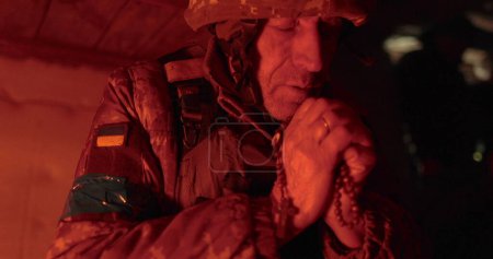 Foto de Retrato cinematográfico de cerca de un viejo soldado orando con esperanza mientras estaba sentado en un foso. Rezando con un rosario en uniforme militar de camuflaje, esperando paz y libertad - Imagen libre de derechos