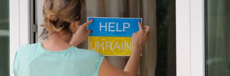 Woman sticking sign on door help Ukraine closeup. Charitable assistance for Ukrainian people concept