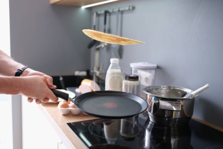 Foto de Hands hold black iron pan and toss pancake into air. Cooking pancakes at home in kitchen - Imagen libre de derechos