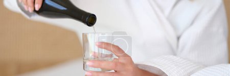 Photo for Woman in bathrobe on bed pours water or white wine into glass alone. Glass of water in morning on empty stomach and alcoholism alone concept - Royalty Free Image