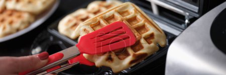 Téléchargez les photos : Pastry chef takes hot waffles from waffle iron in kitchen. Cooking sweet delicious dessert at home - en image libre de droit