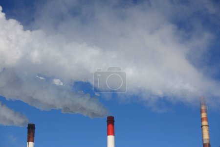 Three pipes of thermal power plant with steam and smoke against blue sky during heating season. Work CHP concept