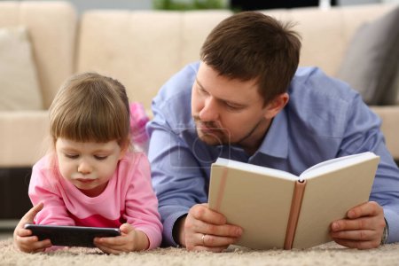 Téléchargez les photos : Plus de communication dans la famille et la dépendance des enfants sur les gadgets. Papa tenant livre enfant regarde l'écran du smartphone - en image libre de droit