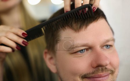 Téléchargez les photos : Coiffeur coupe les cheveux avec des ciseaux et peigne noir. Satisfait client homme assis dans salon de coiffure - en image libre de droit