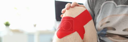 Foto de Male hands hold sore knee with kinesio tape sticker closeup. Injuries and sprains of knee joint and treatment - Imagen libre de derechos