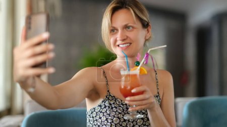 Téléchargez les photos : Sourire femme heureuse tenant cocktail prend selfie prise sur téléphone mobile. Application photo mobile - en image libre de droit