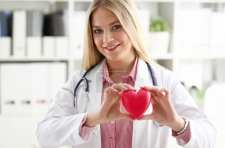 Hermosa sonriente rubia doctora sostener en brazos rojo juguete corazón primer plano. Cardioterapeuta estudiante educación RCP 911 médico salvavidas hacer cardiaca ritmo de pulso físico medida arritmia
