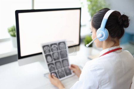 Foto de La doctora se sienta en el consultorio médico en auriculares con micrófono en la computadora y mira los rayos X. Consultas médicas online - Imagen libre de derechos
