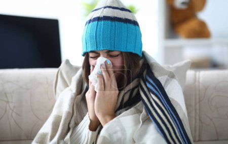 Portrait of beautiful woman blowing into white tissue and closing cute eyes. Poor girl sitting indoors wearing warm clothes and sneezing from severe flu. Hard virus concept