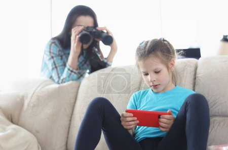 Foto de Mamá controla a su hija con el teléfono a través de prismáticos. Los mejores controles parentales y el concepto de aplicaciones para teléfonos inteligentes - Imagen libre de derechos