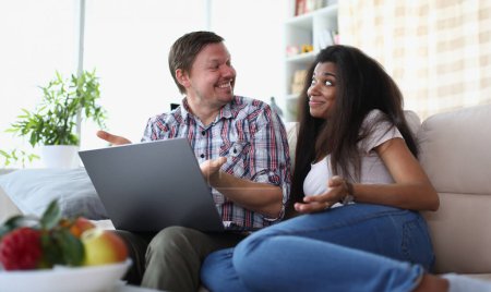 Photo for Interethnic family couple laughing and chatting while sitting on sofa at home with laptop. Married couple making online payments on laptop using financial banking app on computer at home - Royalty Free Image