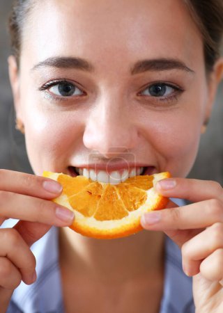 Foto de Mujer retrato sostiene en su mano una rebanada de naranja picada se la come para el desayuno con la boca en la cocina. Concepto de una dieta saludable. - Imagen libre de derechos