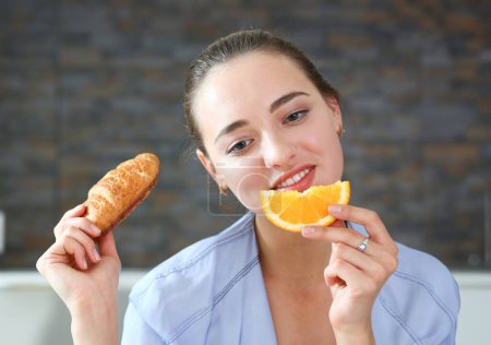 Foto de Mujer retrato sostiene en su mano una rebanada de naranja picada se la come para el desayuno con la boca en la cocina. Concepto de una dieta saludable. - Imagen libre de derechos