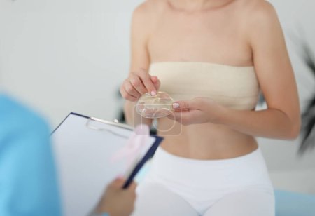 Frau beim Arzttermin hält Silikonimplantat in der Brust. Chirurgisches Konzept zur Brustvergrößerung