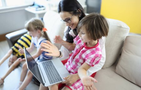 Photo for Woman is sitting on the couch, girl is sitting on her lap, they are looking at laptop next to children are playing. Education quarantined children online concept - Royalty Free Image