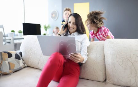 Photo for Woman is sitting on couch holding laptop and talking on smartphone next to the children are playing. Remote work in coronavirus in family concept - Royalty Free Image