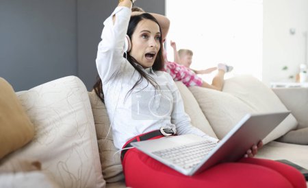 Photo for Woman in headphones and with laptops in her hands sits on sofa from behind, children play. Remote work in coronavirus in family concept - Royalty Free Image
