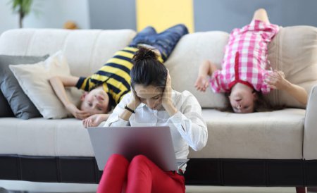 Photo for Woman sits on floor with a laptop and covers her ears with her hands. behind her on couch are children playing around. Remote work at home with children concept - Royalty Free Image