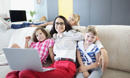 Photo for Woman is sitting on sofa with two girls and a boy is standing next to laptop. Teaching children online in coronavirus concept - Royalty Free Image