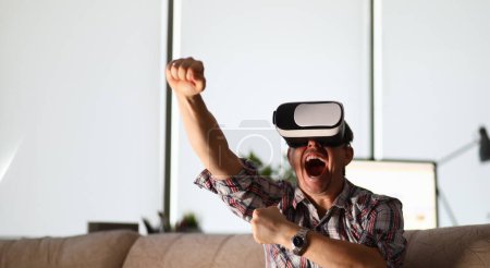 Photo for Attractive man sitting on sofa wearing virtual reality helmet against home background portrait. Battle tournament games remotely concept - Royalty Free Image